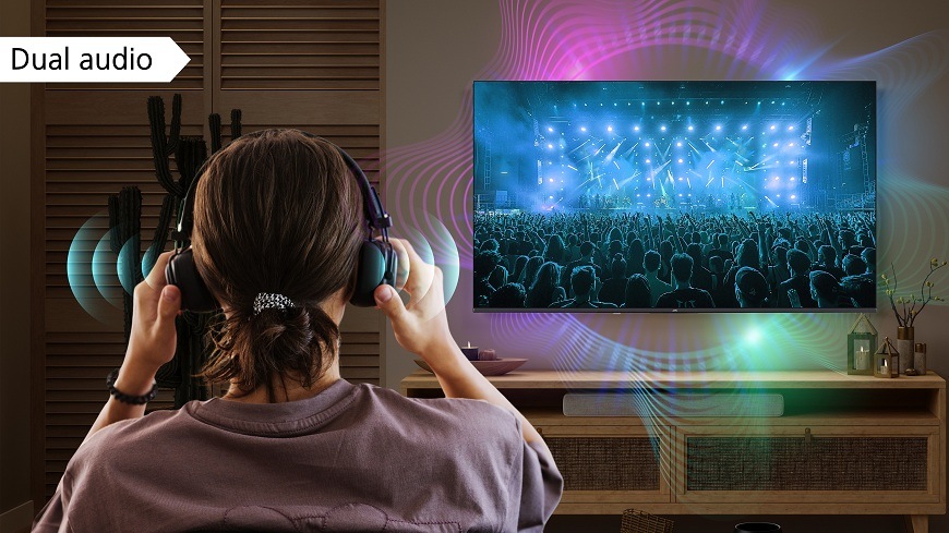 Žena zezadu sedí v obývacím pokoji a má na uších bezdrátová sluchátka. Sleduje JVC TV, na které je dynamický záběr z živého koncertu s mnoha lidmi a jevištními světly. Z TV i ze sluchátek se šíří stylizované zvukové vlny. V levém horním rohu je textová bublina s nápisem "Dual audio". Obrázek ilustruje možnost souběžného poslechu zvuku z TV přes sluchátka i reproduktory