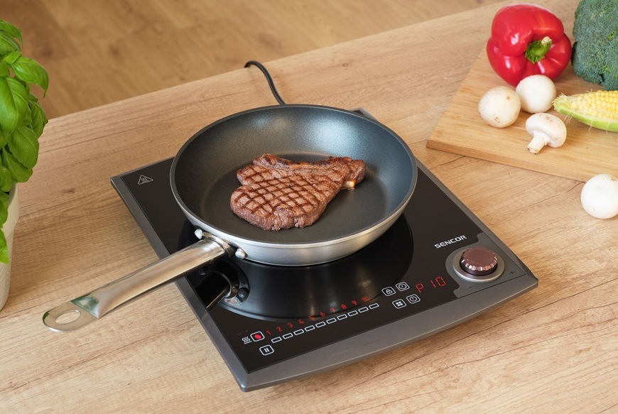 Indukční vařič Sencor SCP 3414GY s dotykovým ovládáním a otočným regulátorem připravuje steak na pánvi na světlé dřevěné kuchyňské lince zatímco v pozadí jsou čerstvé suroviny jako paprika brokolice kukuřice a žampiony na prkénku.