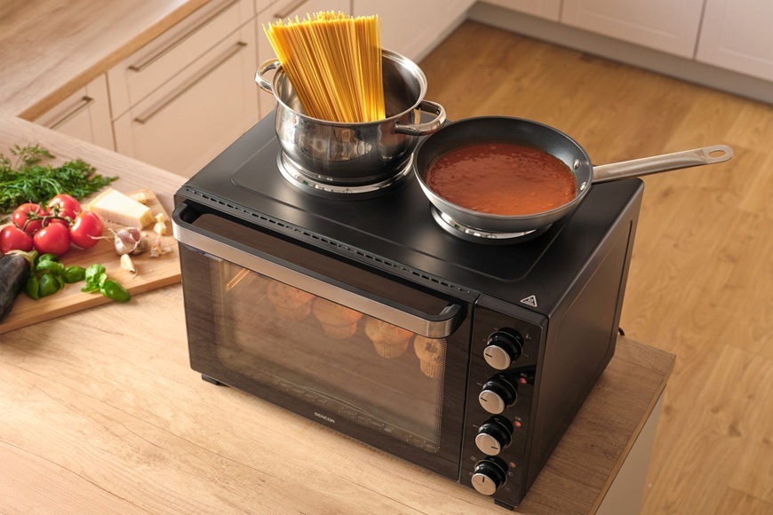 Mini trouba Sencor SEO 4800BK v moderní kuchyni s hrncem špaget a pánví s rajčatovou omáčkou na horní varné ploše, uvnitř se pečou muffiny a kolem jsou čerstvé suroviny na vaření.