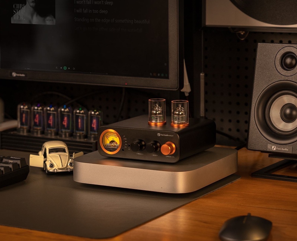 Zesilovač Fosi Audio MC331 černý Na obrázku je zesilovač Fosi Audio MC331 umístěný v pracovně nebo domácím poslechovém koutku. Stojí na stole na stříbrném podstavci, má černé tělo, dvě elektronky na horní straně a výrazný oranžově podsvícený VU měřák i hlavní ovladač na předním panelu. Vpravo vedle něj je reproduktor, vlevo monitor a další drobná elektronika, takže celá scéna působí jako stylově zařízené místo pro poslech hudby. Nahoře je anglický reklamní text, který zdůrazňuje kompaktní rozměry a schopnost zapadnout do různých audio sestav.