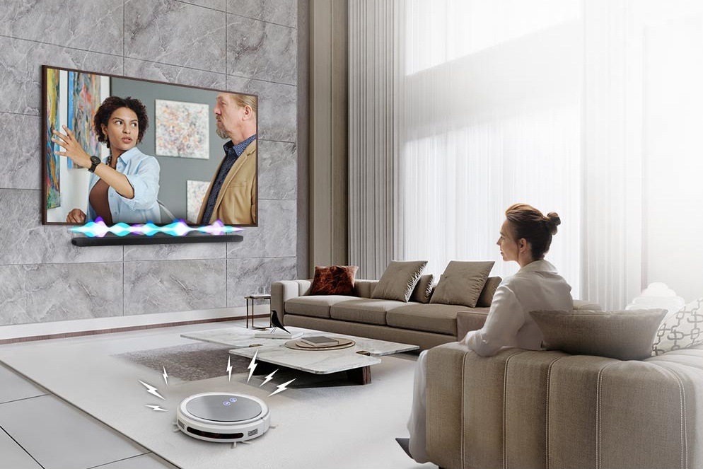 Soundbar Samsung je připevněn na zdi pod televizí Samsung, na jejíž obrazovce spolu hovoří muž a žena, nad soundbarem je nákres proměnlivé intenzity zvuku, v popředí je žena sledující televizi na pohovce a vlevo od pohovky je robotický vysavač