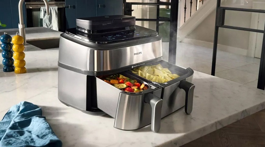 Fritéza horkovzdušná Philips Airfryer Dual Basket 5000 series 9 l, NA555/00 stříbrná Horkovzdušná fritéza Philips se dvěma vysunutými koši umístěná na kuchyňské lince, v jednom koši je připravovaná zelenina a ve druhém brambory, z nichž stoupá pára.