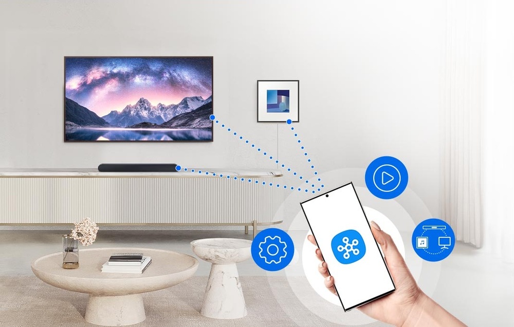 Soundbar Samsung HW-S61D bílý Ruka drží mobil s logem aplikace SmartThings na displeji, kolem mobilu jsou modré ikonky funkcí, na pozadí je místnost s televizí na stěně a soundbarem na televizním stolku, na zdi je reproduktor Music Frame, z mobilu vycházejí modré tečkované linky spojující ho se soundbarem, televizí a reproduktorem