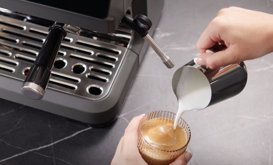 Espresso Hisense HSACM20LP antracitový ženská ruka přeléva našlehane mleko z konvičky do připravené kávy v kávovaru Hisense HSACM20LP