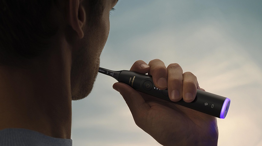 Muž používá elektrický zubní kartáček Philips Sonicare 6500 HX7411/10 při čištění zubů, drží černý kartáček s podsvícenou rukojetí.