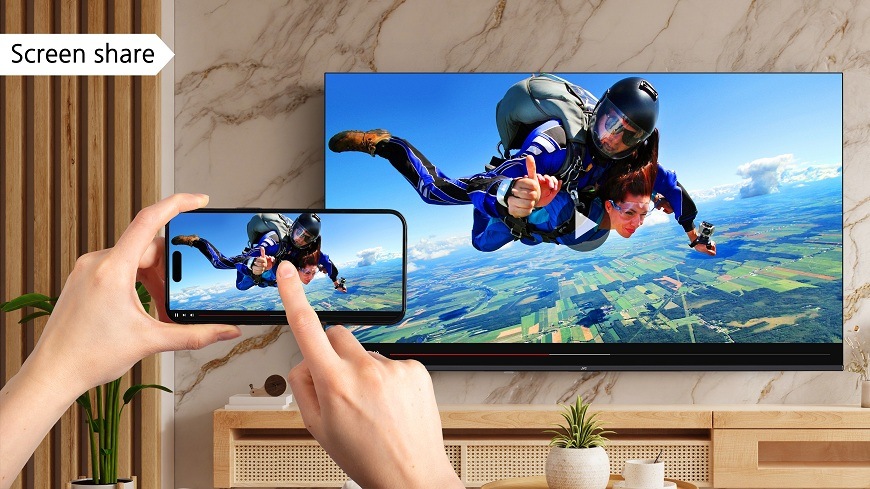 Televize JVC LT-65VDQ9435 Televize JVC ve světlém interiéru, zobrazující funkci sdílení obrazovky. Ruce drží telefon zobrazující seskok padákem. Stejný obraz je bezdrátově sdílen na velké televizi JVC