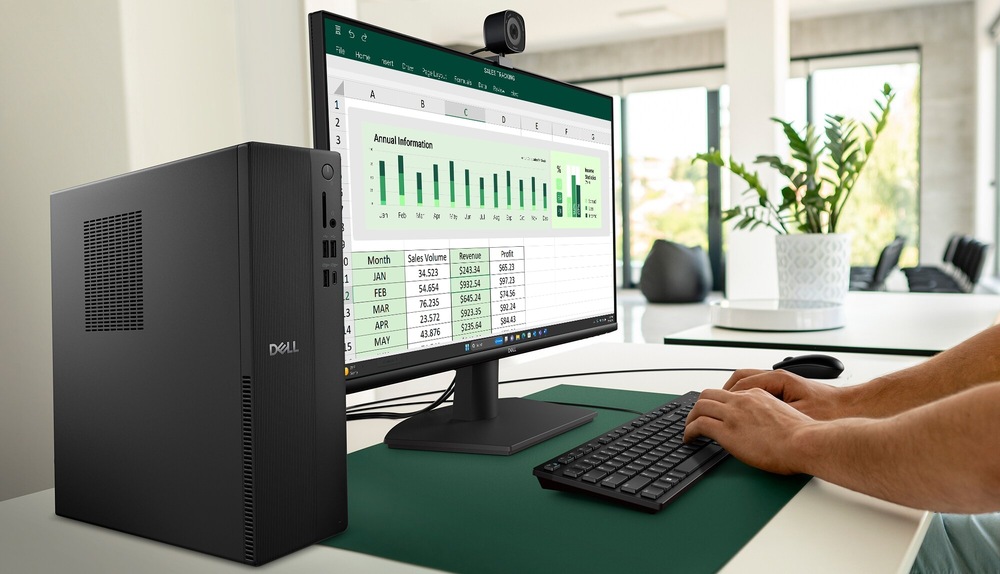 Osoba používá počítač s velkým monitorem, na kterém je zobrazena tabulka Excel s různými datovými grafy a diagramy. Pracovní prostor zahrnuje klávesnici a myš, v pozadí je vidět rostlina.