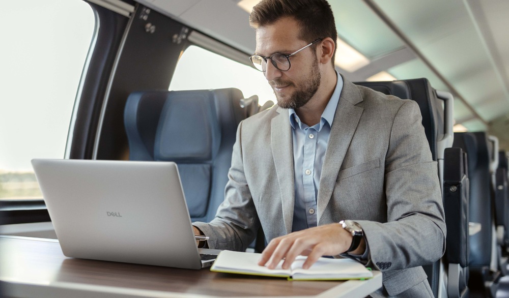 Notebook Dell Pro 16 PC16255 šedý Muž v obleku pracuje na notebooku u stolu ve vlaku. Vypadá soustředěně, má otevřené dokumenty. Interiér vlaku je moderní a dobře osvětlený.