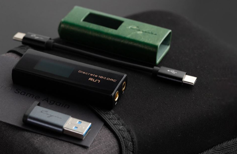 Sluchátkový zesilovač Cayin RU7 černý Sluchátkový zesilovač ležící na látkovém povrchu společně s ochranným pouzdrem a USB adaptérem.