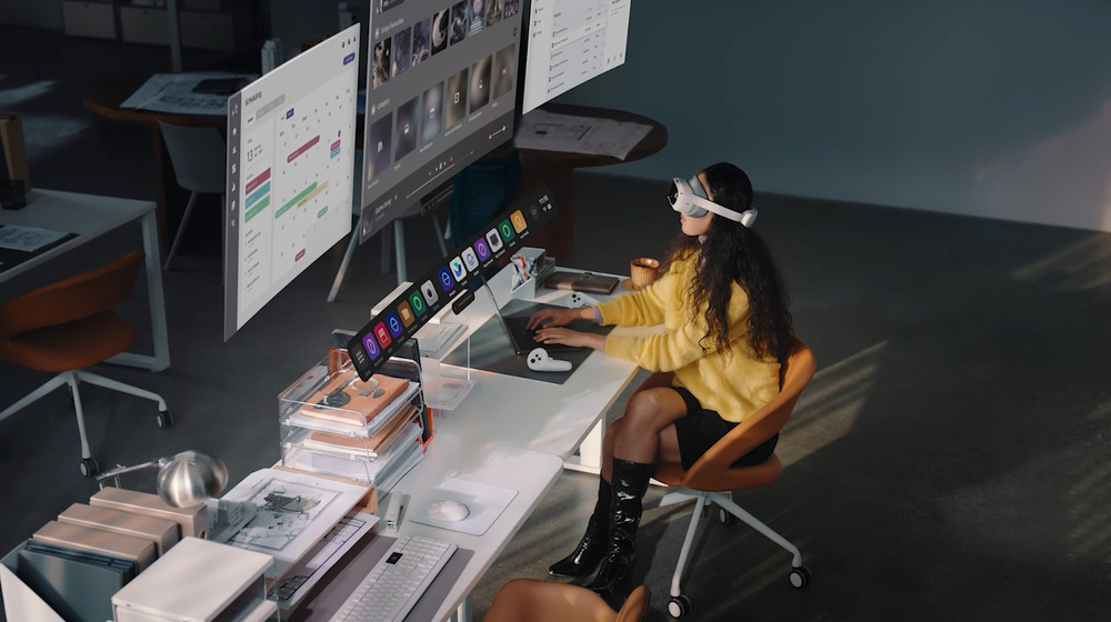  Žena ve žlutém svetru s nasazeným VR headsetem sedí u moderního stolu a interaguje s plovoucími digitálními obrazovkami. Scéna evokuje futuristický pracovní prostor.