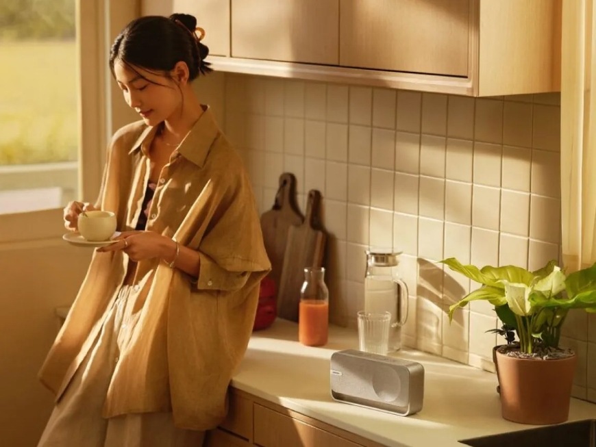 Přenosný reproduktor Bose Soundlink HOME hnědý Žena se šálkem kávy v ruce se opírá o kuchyňskou linku, na které je reproduktor Bose SoundLink Home