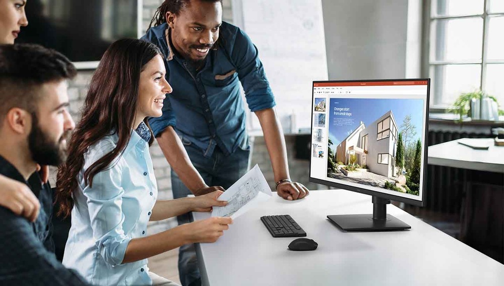 Různorodá skupina kolegů, kteří se usmívají a dívají na monitor zobrazující architektonické návrhy. Světlé, moderní kancelářské prostředí.