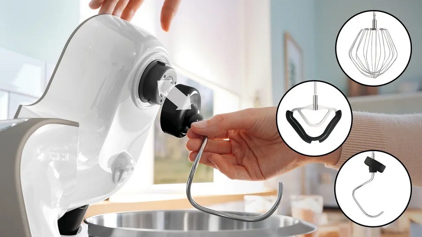 Detailní záběr ruky, která připevňuje hnětací hák do hlavy bílého kuchyňského robotu.Bosch MUM MUM5XL74. V pravé části jsou v kruzích ukázány další nástavce