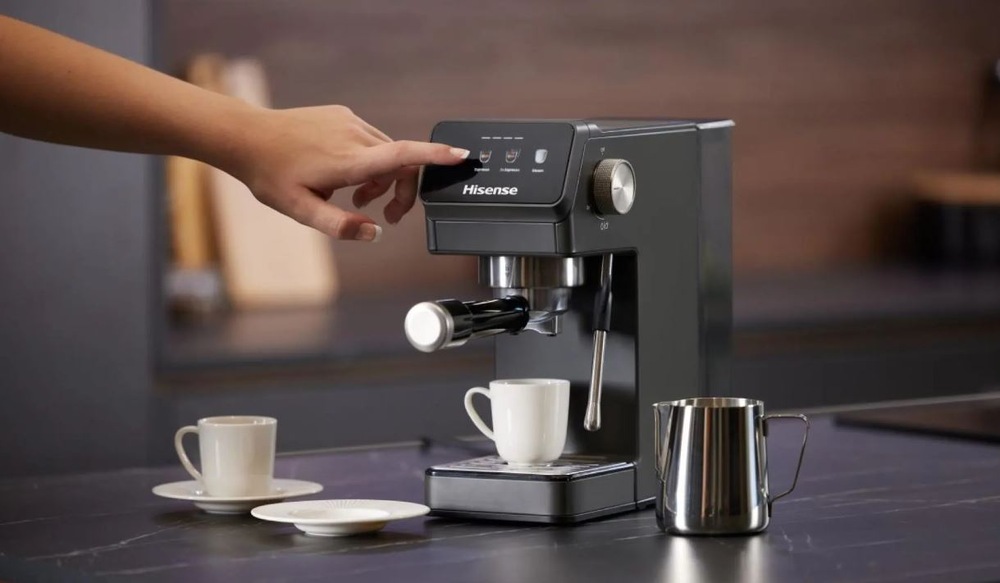 Espresso Hisense HESCM20LP antracitový ženská ruka vybíra program na pákovém kávovaru Hisense HESCM20LP. Vedle kávovaru jsou hrnečky a nádobka na šlehání mléka.