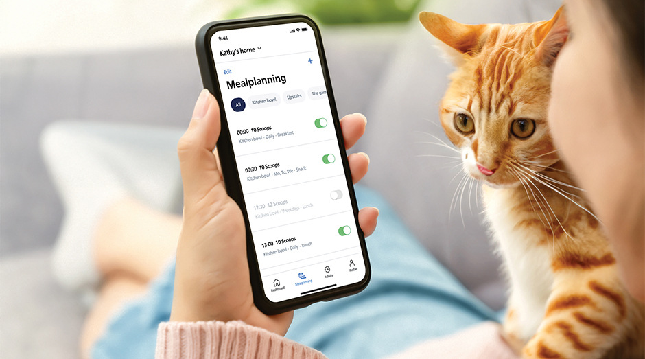 Mobilní aplikace pro chytré krmítko zobrazená na telefonu a kočka sedící vedle osoby, která se na telefon dívá