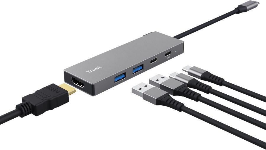 USB Hub Trust DALYX 5v1 stříbrný Na obrázku je stříbrný USB-C hub Trust DALYX 5v1 zobrazený při použití s více kabely. Z pravé strany z něj vede pevný kabel zakončený USB-C konektorem pro připojení k notebooku. Na těle hubu jsou vidět dva modré USB porty, dva menší USB-C porty a na čele HDMI konektor, do kterého je zapojen HDMI kabel. Vedle hubu leží ještě další USB a USB-C kabely, takže obrázek zdůrazňuje možnosti připojení více zařízení najednou.