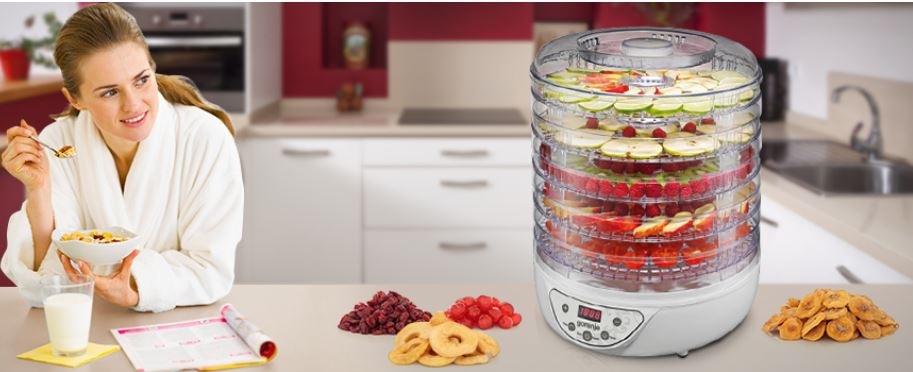 Sušička ovoce Gorenje FDK 24 DW bílá/plast žena v županu snídá kaši z misky, v níž má usušené ovoce, které sušila v sušičce na stole vedle ní. Sušička je plná potravin a vedle ní jsou další suroviny.