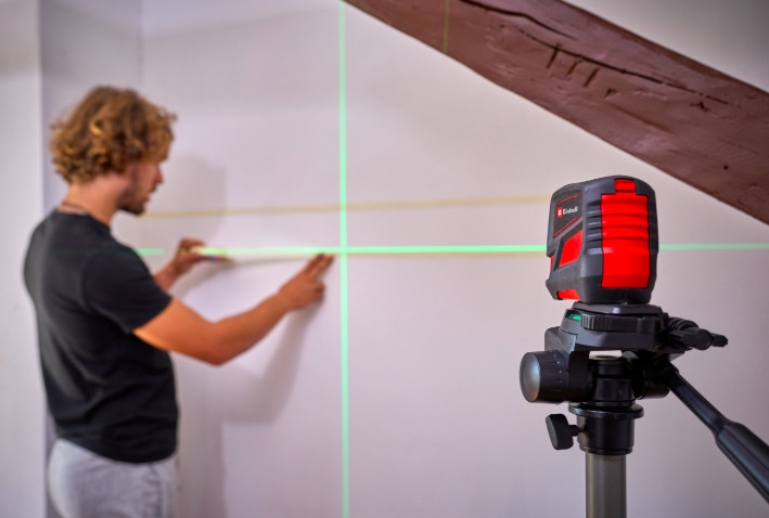 Muž používá křížový laser Einhell TCLL2G na stativu při zarovnání poličky na stěně