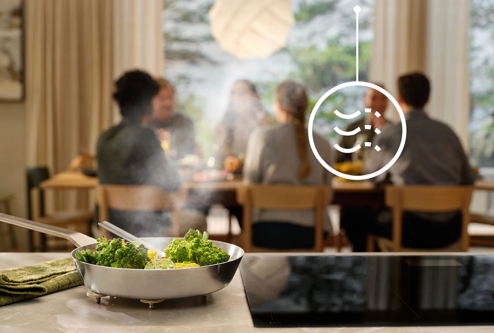 Odsavač par Electrolux Série 700 LFV449K černý V popředí je indukce Electrolux. Vedle je položená pánev se zeleninou. V pozadí sedí lidé u stolu. Nad deskou je symbol pro funkci Breeze.