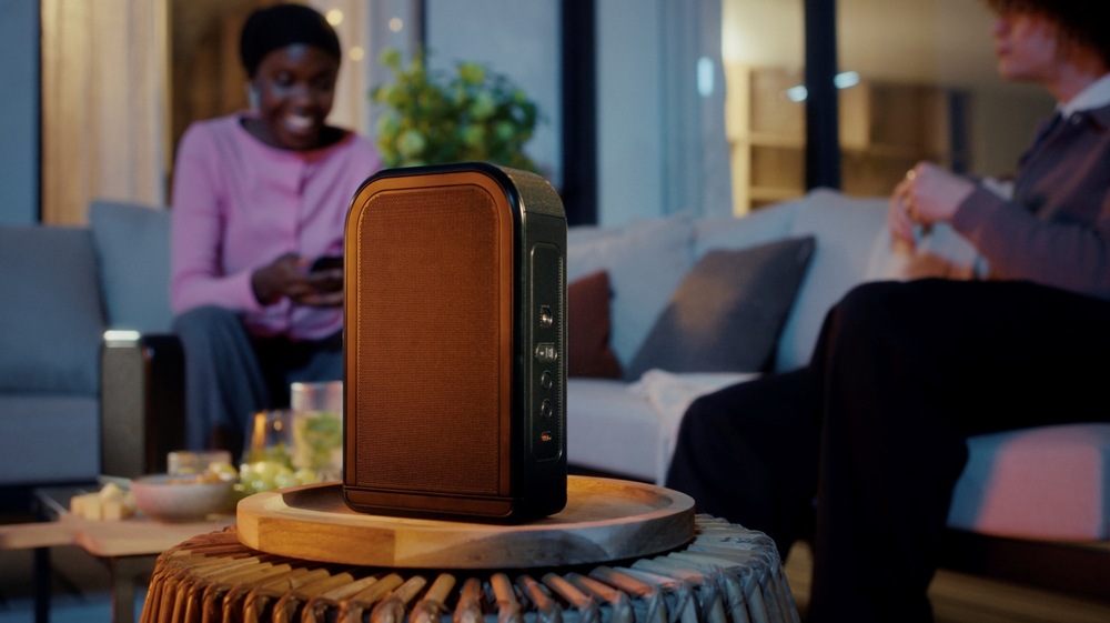 Dva lidé si povídají v obývacím pokoji, žena drží mobilní telefon, v popředí je odnímatelný bezdrátový reproduktor soundbaru JBL BAR 1300 MK2 na stolku