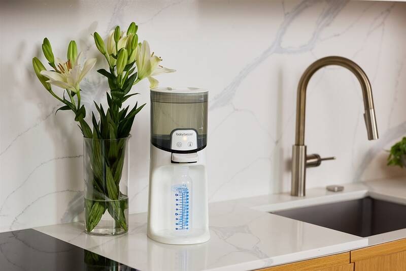 Ohřívač vody Baby Brezza Instant Warmer White bílý Ohřívač vody stojí vedle kuchyňského dřezu, těsně za přístrojem se nachází kytice ve váze.