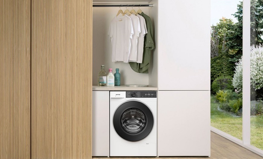 Pračka Gorenje G400 W484A3 UltraClean bílá pračka Gorenje W484A3 UltraClean vedle dřevěné šatní skříně. Nad pračkou visí na ramínkách oblečení. Na prave straně jsou velké prosklené dveře do zahrady.
