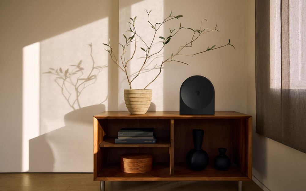 Reproduktory Samsung Music Studio 5 LS50H (2 ks) černé Minimalistický interiér s černým reproduktorem Samsung HW-LS50H Studio 5 na dřevěné skříňce vedle květináče s větvemi, osvětlený teplým slunečním světlem vrhajícím stíny na zeď.