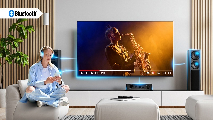 Televize JVC LT-65VDQ9435 Žena se sluchátky sleduje hudební video na svém telefonu v moderním interiéru, stejný hudební obsah se promítá na televizi JVC, což demonstruje bezdrátové Bluetooth připojení zařízení k JVC TV