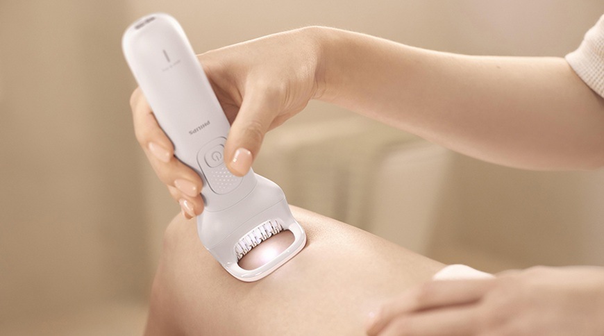 Epilátor Philips BRE750/00 v bílé barvě přiložený k pokožce nohy, detail záběru na epilační hlavici s osvětlením při odstraňování chloupků.