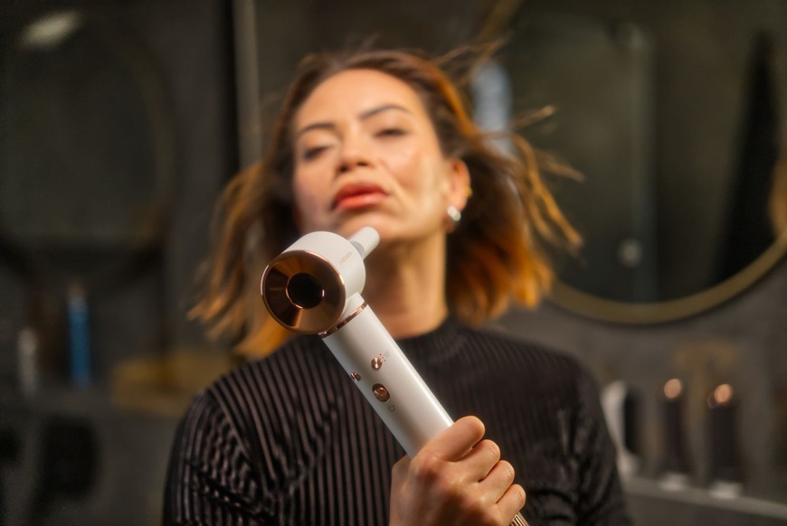 Žena s krátkými vlasy používá foukací kulmu Sencor SHS 9100RS Artemiss, kterou drží v ruce při úpravě účesu. Proud vzduchu z kulmy zvedá prameny vlasů, zatímco pozadí tvoří interiér koupelny se zrcadlem a kosmetickými doplňky.