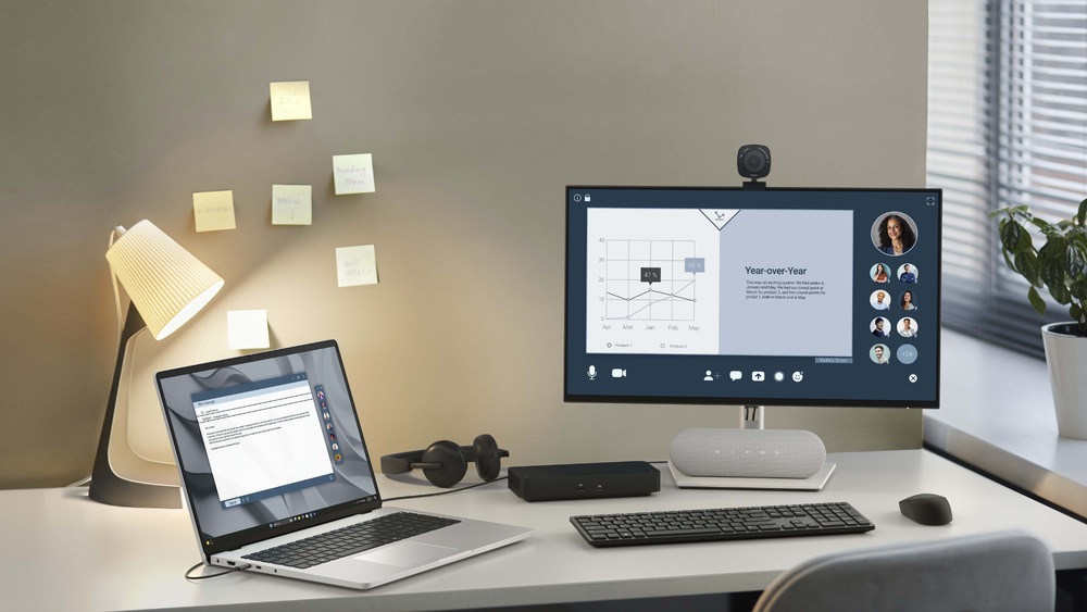 Notebook Dell Pro 16 PC16255 šedý Moderní pracovní stůl s notebookem a na desce stolu umístěným monitorem, na kterém se zobrazují grafy a rozhraní pro videohovory. Na zdi jsou nalepené poznámkové lístky, lampička a zelené rostliny dodávají místnosti útulný vzhled.