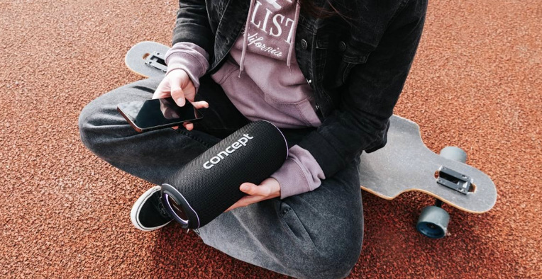 Osoba sedící u skateboardu drží reproduktor Concept SoundJoy BR5530 a ovládá hudbu telefonem