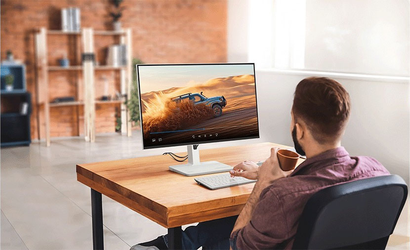 Monitor Lenovo L27h-4A šedý Muž sedí u dřevěného stolu v moderní kanceláři, drží hrnek s kávou a na obrazovce počítače sleduje terénní vozidlo řítící se pískem.