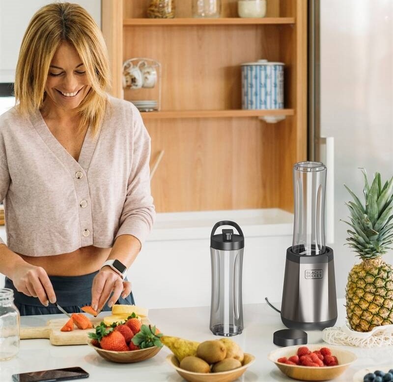 Stolní mixér Black+Decker BXJB1204E TO-GO nerez Usměvavá žena připravuje na kuchyňské lince ovoce pro čerstvé smoothie, vedle toho je umístěn mixér Black+Decker BXJB1204E