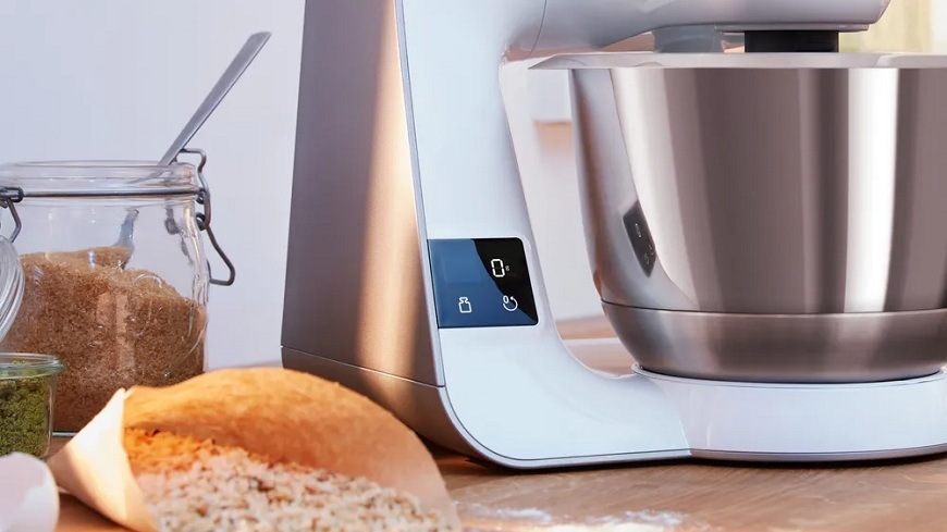 Detail kuchyňského robota Bosch MUM5XW00 s nerezovou mísou a dotykovým panelem, umístěného na kuchyňské lince vedle sklenic se surovinami, papírového sáčku se sypkými ingrediencemi a lžíce připravené na pečen