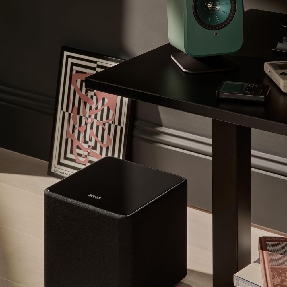 Subwoofer KEF Kube 8 MIE (1 ks) černý Černý subwoofer KEF Kube 8 MIE umístěný na podlaze vedle stolku se zeleným reproduktorem KEF a přenosnými přehrávači.