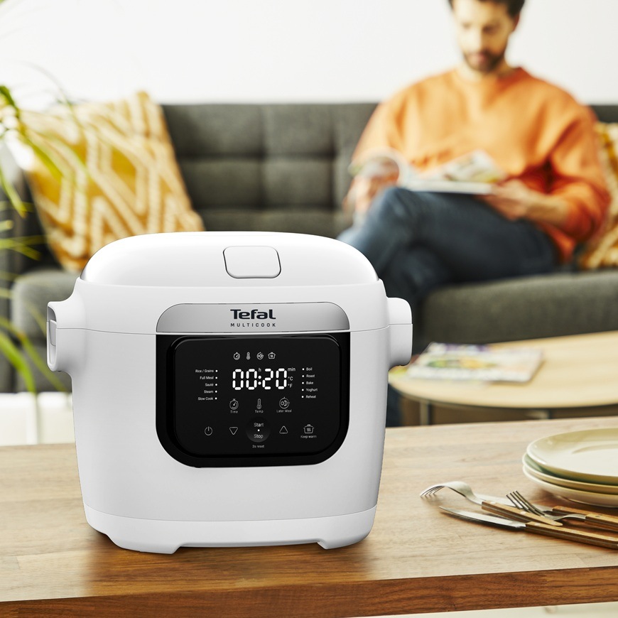Multifunkční hrnec Tefal MY700BF0 Multicook s digitálním displejem stojí na stole v obývacím pokoji, zatímco muž v pozadí čte časopis na pohovce.