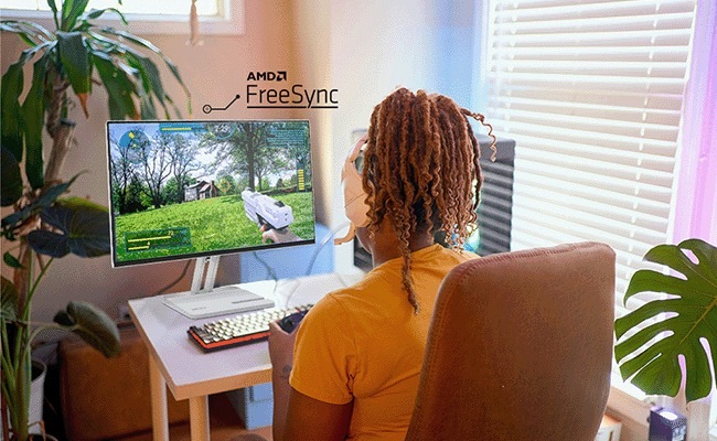 Monitor Lenovo L24-4C šedý Osoba sleduje videohru na monitoru se zapnutou technologií AMD FreeSync. Sedí v útulné místnosti s rostlinami a slunečním světlem proudícím dovnitř.