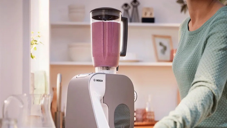 Mixér naplněný koktejlem připojený na kuchyňský robot Bosch MUM5XL74, na pravé straně je vidět část ženské postavy obsluhující mixér
