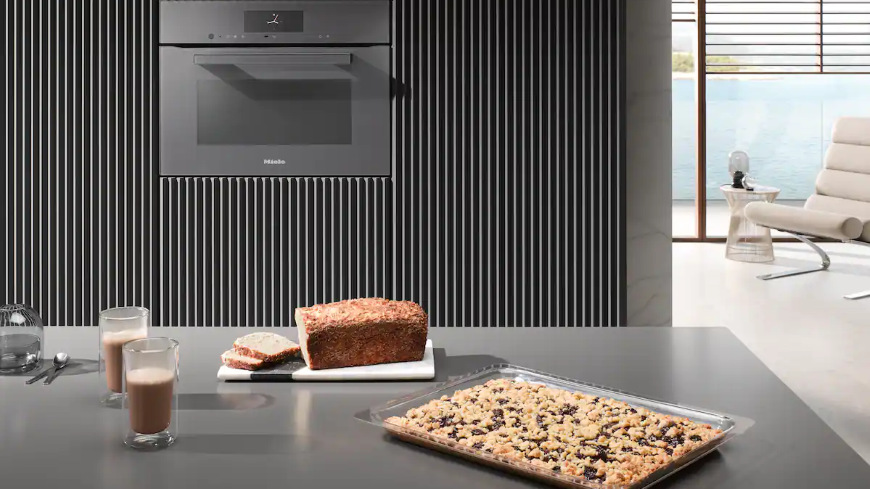 Trouba Miele H 7440 BM béžová Pekáč s koláčem položený na pracovní desce, za ním upečený chléb a v pozadí trouba Miele H 7440 BM