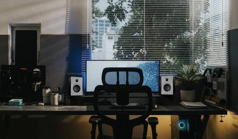 Herní setup s reproduktory Swan M10 Plus u monitoru, s podsvícením PC a výhledem z okna se žaluziemi.