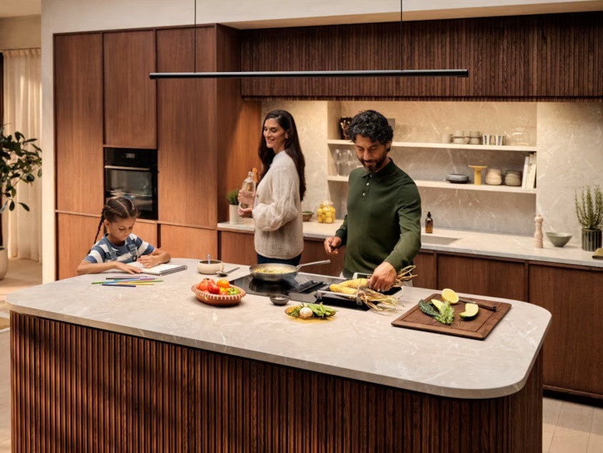 Indukční varná deska s odsáváním Electrolux Série 600 ECC8344 černá Stylová rodinná kuchyně s varnou deskou s integrovaným odsáváním, která uprostřed kuchyňského ostrůvku zajišťuje čistý vzduch přímo při vaření. Muž připravuje čerstvé ingredience na elegantní varné ploše, zatímco dítě si u stejné pracovní desky kreslí a žena v pozadí podává vodu.