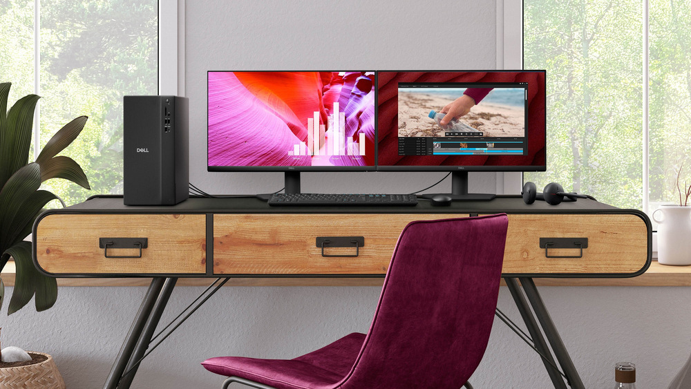 Stolní počítač Dell Pro Tower Essential QVT1260 černý Moderní pracovní prostor s dřevěným stolem, dvěma monitory, stolním počítačem a sluchátky na boku. Před stolem stojí růžová židle a z oken je vidět zeleň.