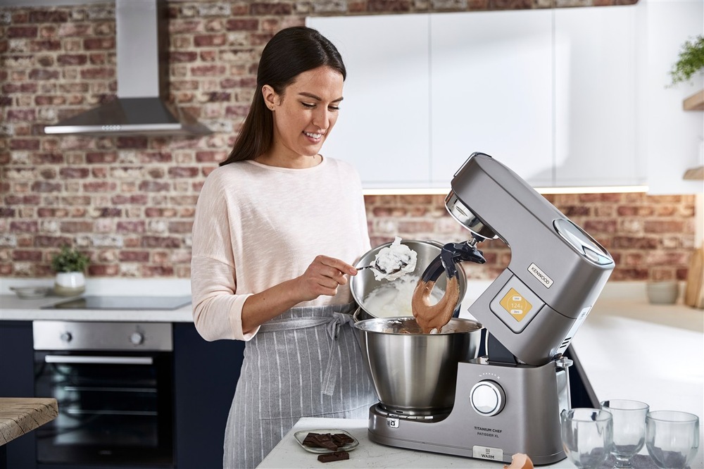 Kuchyňský robot Kenwood KWL90.594SI se zvednutým ramenem stojí na kuchyňském stole, žena přidává do mísy robota z nádoby v ruce suroviny