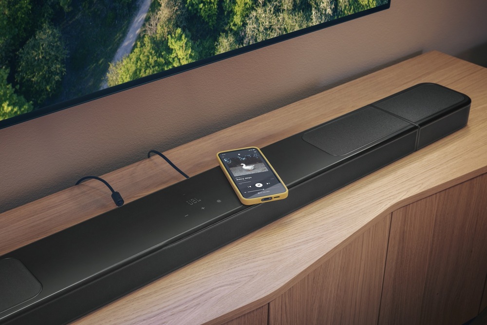 Soundbar JBL BAR 1300 MK2 položený na televizním stolku, na soundbaru leží mobilní telefon