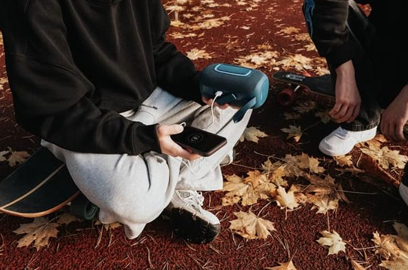 Přenosný reproduktor Concept SoundJoy BR5022 modrý Mladý muž dobíjí telefon z modrého reproduktoru Concept SoundJoy BR5022 při venkovním posezení