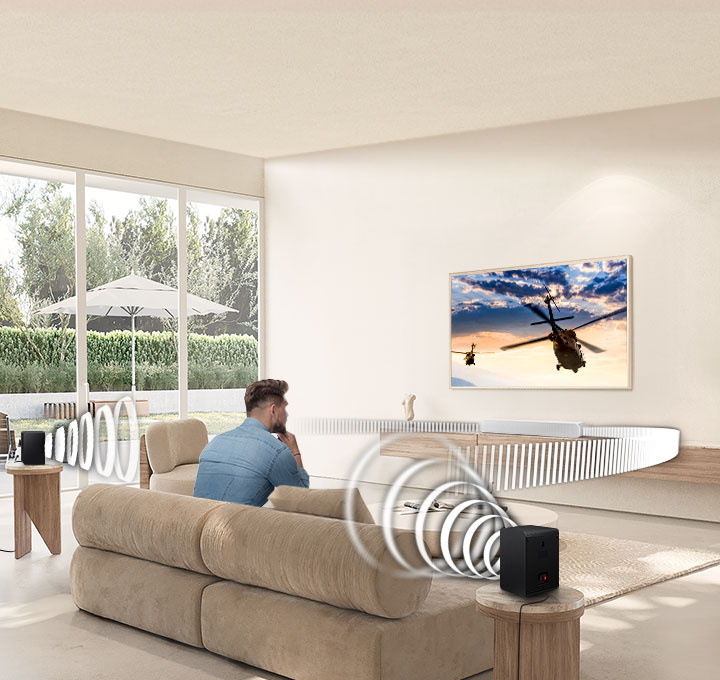 Soundbar Samsung HW-S61D bílý Muž na gauči sleduje televizi, z reproduktorů kolem něj a soundbaru Samsung ležícího pod TV vychází zvuk znázorněný bílými čárami