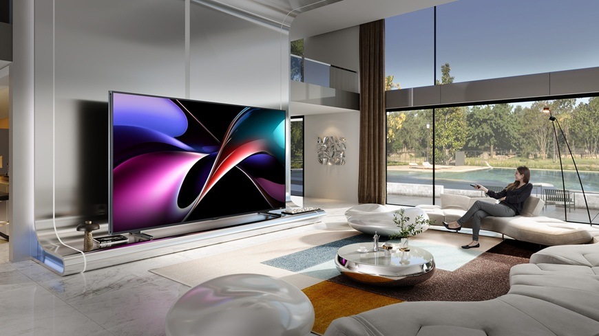 Televize Hisense 116UXQ Na obrázku je žena, sedící na gauči a zapíná televizor Hisense 116UXQ pomocí dálkového ovladače
