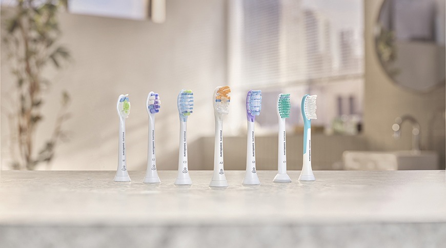 Různé náhradní hlavice pro elektrické zubní kartáčky Philips Sonicare uspořádané v řadě na koupelnové desce, každá s jiným typem štětin.