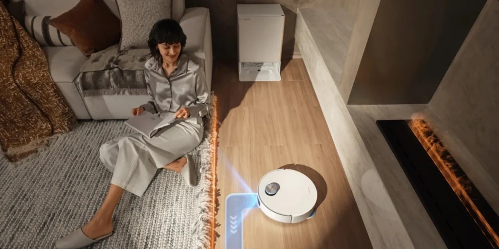 Vlevo mobil s uživatelským rozhraním mobilní aplikace. Vedle něj sedí žena na koberci opřená o pohovku. Vedle koberce vysává podlahu robotický vysavač Dreame Matrix10 Ultra.
